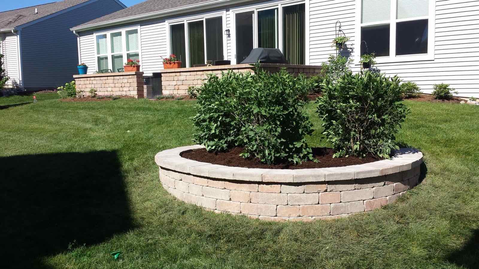 Circular raised stone planter bed with flowering shrubs in a backyard lawn