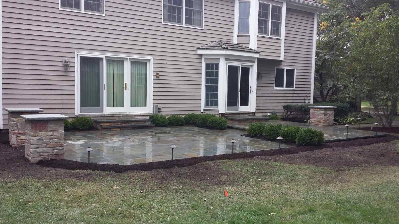 Bluestone patio with stone pillars, landscape lighting, and boxwood hedges